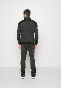 Tuta sportiva nera con giacca con zip e pantaloni tapered. Include strisce bianche sui maniche e sui lati. Texture liscia.