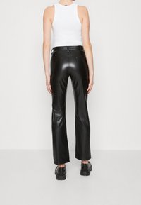 Pantalons évasés en simili cuir noir avec taille haute, deux poches à l'arrière et une texture lisse. Associés à des chaussures noires imposantes.
