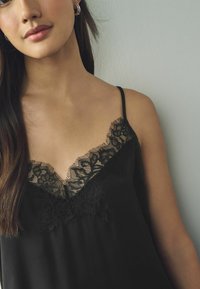 Zwarte camisole met delicate kantdetails langs de V-hals. Gemaakt van zachte stof met dunne verstelbare bandjes.