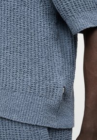 Gros plan sur un haut tricoté bleu texturé à manches courtes et un bas assorti porté par une personne à la peau foncée.