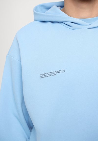Heledad sinine kapuuts, valmistatud orgaanilisest puuvillast. Omab kapuutsi, vaba lõiget ja esit on väike tekst detail kanga töötlemise kohta.