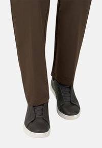 Mustat nahkakengät, joissa on joustavat paneelit ja harmaat nauhat, yhdistettynä ruskeisiin housuihin. Kengissä on valkoinen kumipohja ja teksturoitu muotoilu.