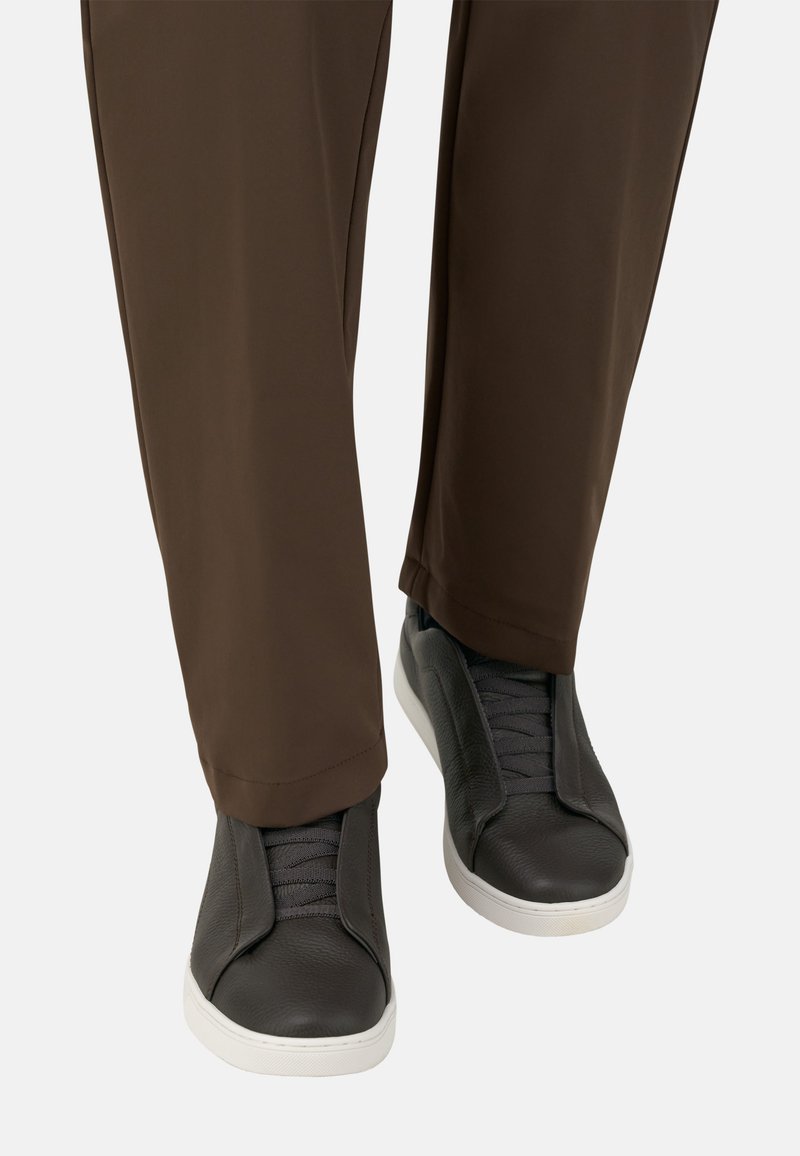 Mustat nahkakengät, joissa on joustavat paneelit ja harmaat nauhat, yhdistettynä ruskeisiin housuihin. Kengissä on valkoinen kumipohja ja teksturoitu muotoilu.