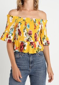 Top amarillo con escote al hombro y patrón floral, corpiño fruncido y mangas con volantes. Combinado con jeans azules de talle alto.