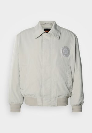 Hellgraue Bomberjacke mit durchgehendem Reißverschluss, gerippten Bündchen und Saum. Verfügt über einen runden Logo-Patch auf der linken Brustseite. Glatte Textur.