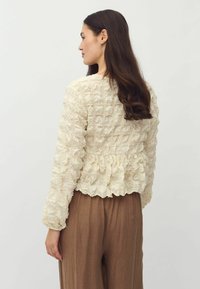 Krémová textúrovaná bunda s volánovým dizajnom, dlhými rukávmi a peplum lemom, v kombinácii s hnedými, voľne strihanými nohavicami.