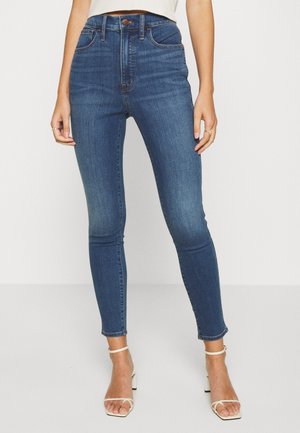 Jeans skinny de cintura alta em denim azul escuro com desgaste discreto. Apresenta cinco bolsos e fecho de botão. Usado em conjunto com sapatos de salto alto com tiras.