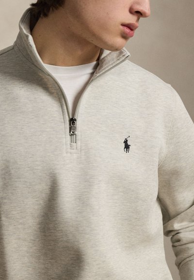Jong persoon draagt een lichtgrijze kwart-ritssluiting trui met een klein donker polo-spelerlogo op de borst, kijkt naar rechts.