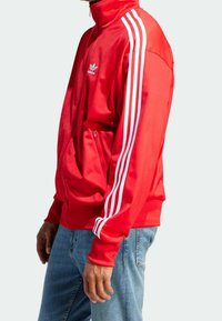 Punainen urheilutakki, jossa on korkea kaulus, valkoiset raidat hihoissa, vetoketjullinen tasku ja sileä kangaspinta.