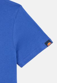 Tricou albastru cu mânecă scurtă, confecționat din bumbac, având o textură netedă și un patch cu logo în contrast pe mânecă.