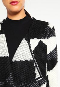 Cardigan bianco e nero con motivo a fantasia, realizzato in tessuto testurizzato, con colletto a scialle e cuciture a contrasto lungo i bordi.