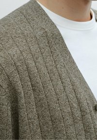 Olivgrön stickad cardigan med ribbad textur, med V-ringning och knapppar. Bärs över en vit skjorta.