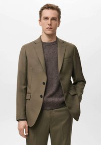 Maslinasto zeleni sako s reverima s usjekom i dva crna gumba, uparen s teksturiranim smeđim džemperom, koji ima uski kroj i glatki materijal.