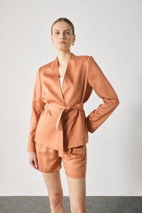Peach satin blazer with a wrap design and belt detail, paired with matching shorts. Smooth texture, long sleeves, and tailored fit.