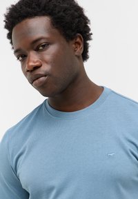 Jasnoniebieski T-shirt z okrągłym dekoltem wykonany z miękkiego materiału, z małym haftowanym logo na piersi. Gładka faktura z krótkimi rękawami.