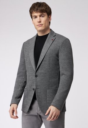 Blazer grigio a righe con revers a notch, chiusura con un solo bottone e due tasche frontali. Indossato sopra un maglione nero; texture liscia.