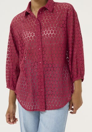 Kobieta w przezroczystej, ciemnoróżowej koronkowej bluzce zapinanej na guziki z okrągłymi wzorami i rękawami do połowy przedramienia, w połączeniu z jasnoniebieskimi dżinsami.