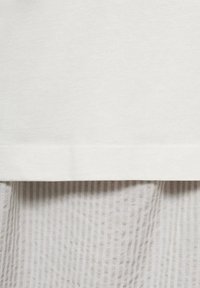 T-shirt bianco in cotone con orlo dritto, sovrapposto a un tessuto grigio chiaro testurizzato con righe verticali.