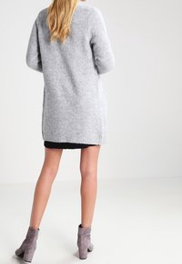 Gråstickad lång cardigan med en mjuk textur och oversized passform. Den har sidofickor och ribbade ärmslut, i kombination med en svart klänning och grå stövlar.