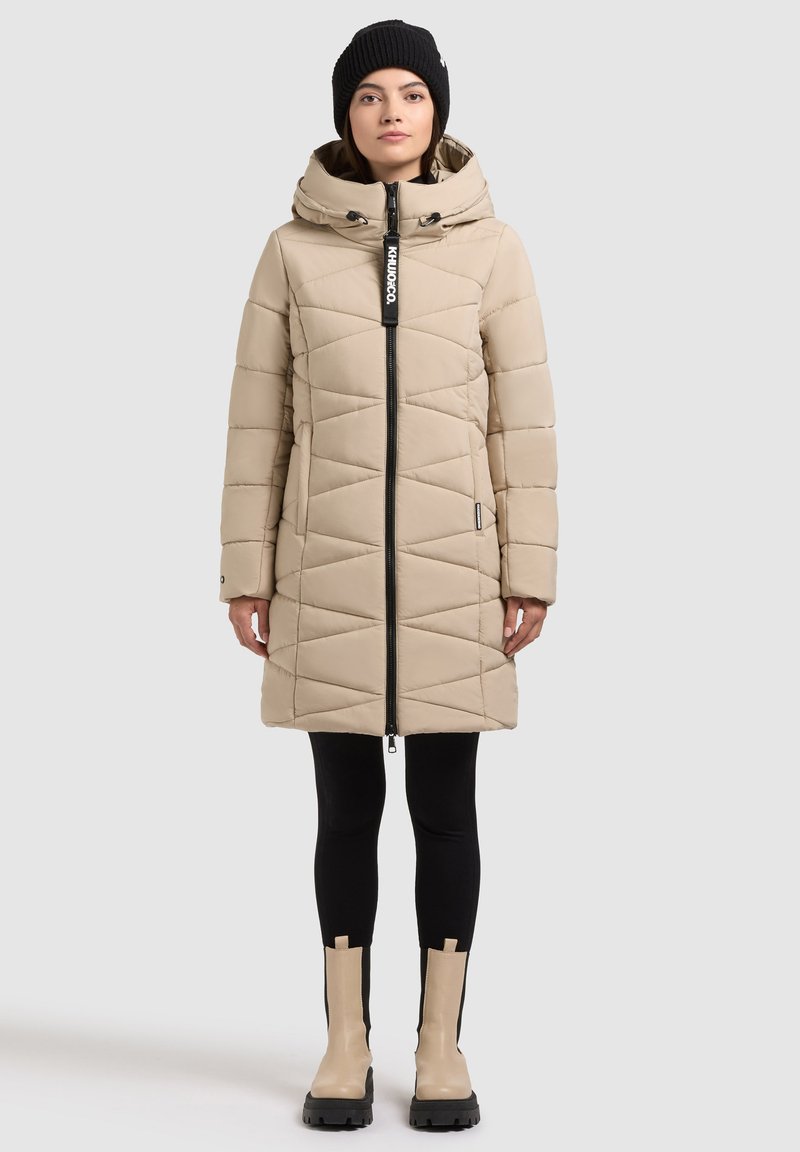 Manteau matelassé beige avec une fermeture éclair à l'avant, une capuche et un motif géométrique matelassé. Porté par-dessus des leggings noirs et des bottines beiges avec des semelles noires.