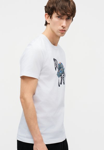 Νεαρός άνδρας που φοράει άσπρο T-shirt με γραφικό μιας ζέβρας που έχει σχεδιασμό αστικού τοπίου στο σώμα της, στέκεται μπροστά σε απλό φόντο.