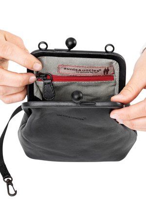 Ręce otwierające czarny skórzany portmonetkę z ramką zatrzaskową, ukazujące wnętrze wyściełane paskowaną tkaniną z kieszonką na zamek w kolorze czerwonym oraz etykietą „aunts & uncles”.