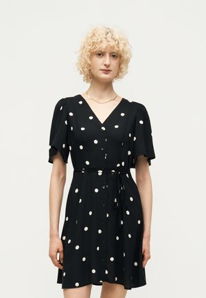 Kvinde med kort, krøllet blondt hår iført en sort kortærmet kjole med hvide prikker, knapper foran og bælte bundet i taljen.