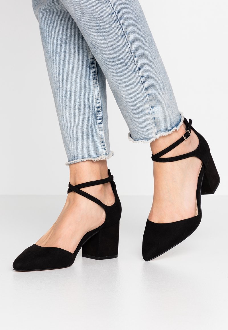 Pieds portant un jean bleu clair à ourlet effiloché et des chaussures à talons blocs en suède noir avec des brides croisées à la cheville.