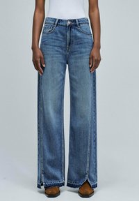 Model wearing high-waist blue wide-leg jeans with front slits at the hem and brown square-toe shoes on a plain background.