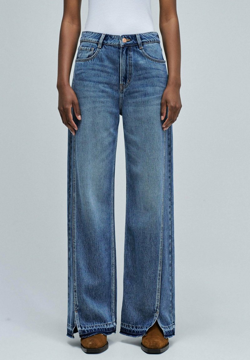 Model wearing high-waist blue wide-leg jeans with front slits at the hem and brown square-toe shoes on a plain background.