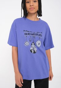 Camiseta de algodón morada con un diseño gráfico que presenta ilustraciones negras de un sol, un símbolo de paz y un reloj. Corte holgado y mangas cortas.