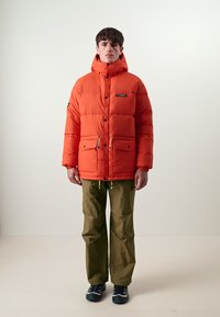Oransje puffjakke med hette og lommer foran, med en svart logo-lapp. Kombinert med olivengrønne bukser og blå sportssko.