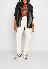 Camisa de poliéster texturizado negro sobre una camiseta gráfica roja con un oso. Pantalones amplios color crema con aberturas laterales y zapatillas blancas.