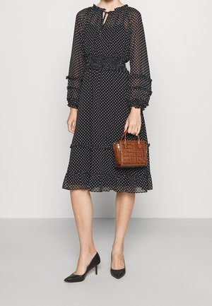 Vestido negro con lunares, de mangas largas y transparentes, cintura fruncida y falda en capas. Bolso marrón con patrón de cocodrilo y tacones negros de punta.