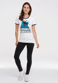 White t-shirt with a blue Cookie Monster graphic and black accents at the collar and sleeves, paired with black skinny jeans and white sneakers.