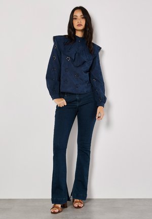Marineblauw blouse met een grote geribbelde kraag, bloemvormige uitsparingen en lange mouwen, gecombineerd met donkerdenim flared jeans en bruine sandalen.