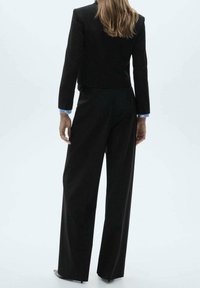 Veste de costume noire ajustée associée à un pantalon à jambes larges. La veste présente une coupe courte, avec des poignets rehaussés d'un accent bleu clair.