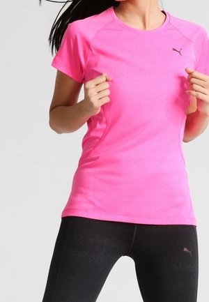 Femme portant un t-shirt de sport à manches courtes Puma rose vif et des leggings noirs Puma, posant avec les mains près de la poitrine.