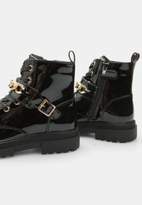 Botine negre din piele la gaură cu detalii din metal auriu, șireturi în față, fermoar pe lateral și talpă chunky din cauciuc. Caracteristici includ accente metalice și detalii cu cataramă.