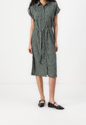 Vrouw draagt een grijze, knielange jurk met knopen, korte mouwen en een zwart-witte luipaardprint, gecombineerd met zwarte platte schoenen met zilveren oogjes.