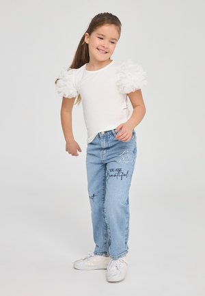 T-shirt bianco con maniche di pizzo arricciate e trasparenti, abbinato a jeans azzurri chiari con dettagli ricamati e scritte. Sneakers bianche e scintillanti.