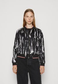 Veste bomber noire avec un motif tie-dye en blanc et rose clair. Elle présente des poignets et un bas côtelés, une fermeture éclair à l'avant, et des accents roses au niveau du col.