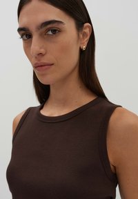 Braunes geripptes Tanktop mit rundem Ausschnitt, das eine taillierte Form zeigt. Kleine vergoldete Ohrringe betonen das Ohr. Neutrale Hintergrund.