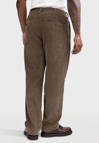 Pantalons en velours côtelé marron avec des côtes verticales, deux poches arrière avec boutons, et une coupe décontractée. Portés avec des chaussures marron foncé.