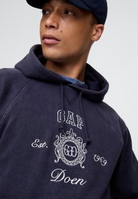 Tengeri kék pamut kapucnis pulóver, elején fehér hímzett "GAP" és "Doen" felirattal. Raglán ujjakkal és húzózsinóros kapucnival.