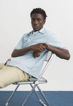 Young man with short curly hair wearing light blue shirt and beige pants, sitting relaxed on a metal chair against plain white background.