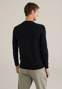 Hombre vestido con un suéter negro de manga larga y pantalones beiges, de pie con la espalda vuelta y la cabeza ligeramente girada hacia la derecha, contra un fondo neutral.