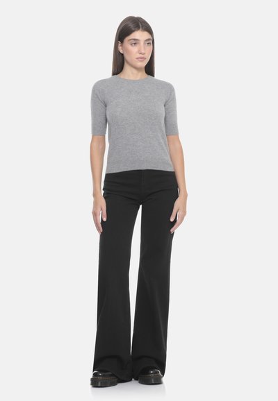 Maglione grigio a maniche corte realizzato in materiale morbido, abbinato a pantaloni a vita alta neri svasati. Il modello è in piedi contro uno sfondo bianco.