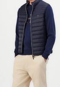 Homme portant un gilet matelassé bleu marine par-dessus un sweat-shirt bleu marine avec logo, pantalon beige, mains dans les poches, debout devant un fond blanc.