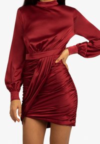 Czerwona satynowa sukienka z długimi bufiastymi rękawami, wysokim dekoltem i fałdowanym wzorem po boku. Posiada dopasowany pas, który tworzy efekt drapowania.